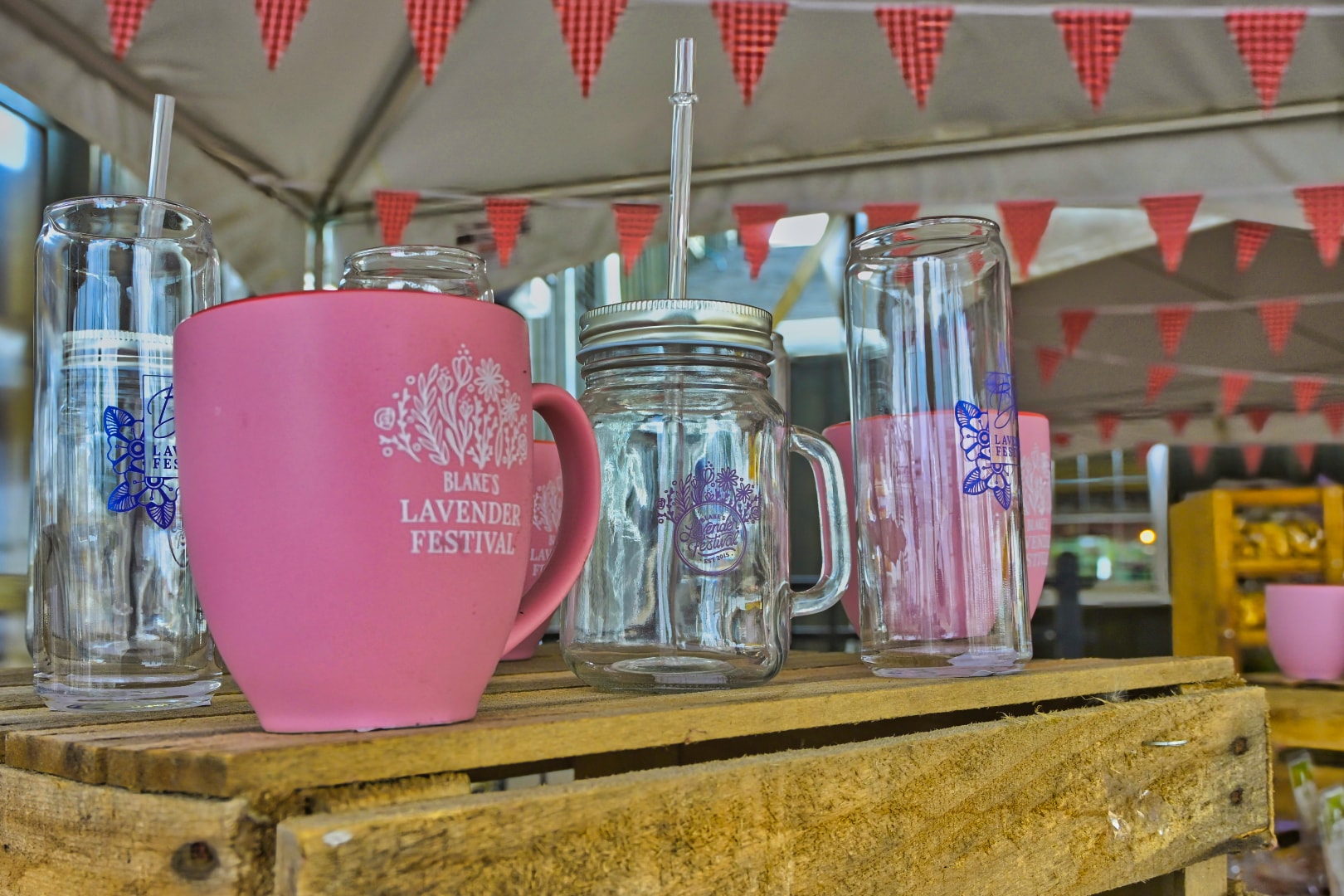 Blake's Lavender Festival branded drinkware display — pink ceramic mug, mason jar, and glass can tumblers with lavender festival logo on a rustic wood pallet shelf under a vendor tent, Armada, Michigan — festival merchandise and event photography by Atelier Lens LLC
