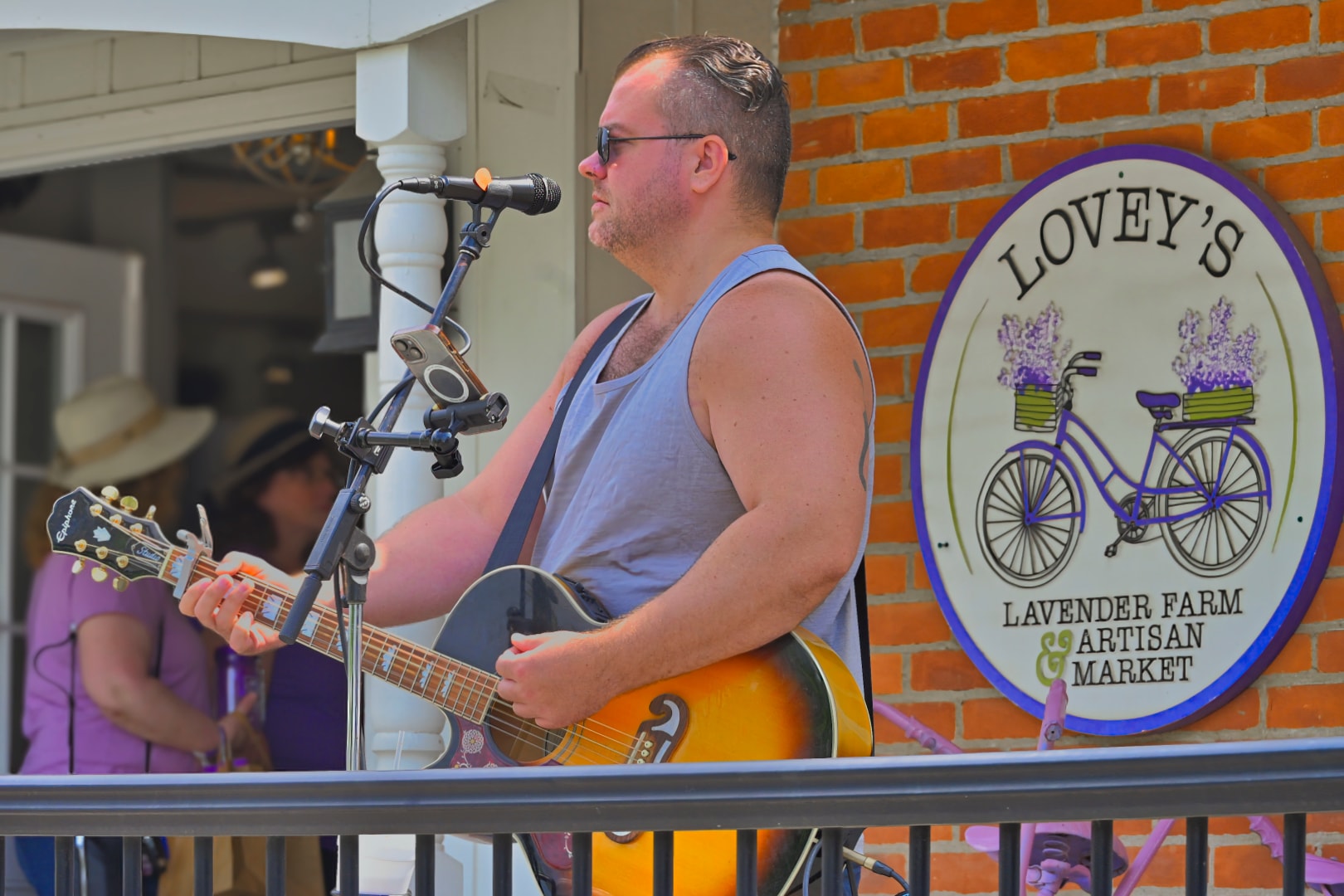Acoustic guitarist performing live on stage in front of the Lovey's Lavender Farm & Artisan Market sign at Blake's Lavender Festival, Blake's Apple Orchard, Armada, Michigan — live music and outdoor festival event photography by Atelier Lens LLC