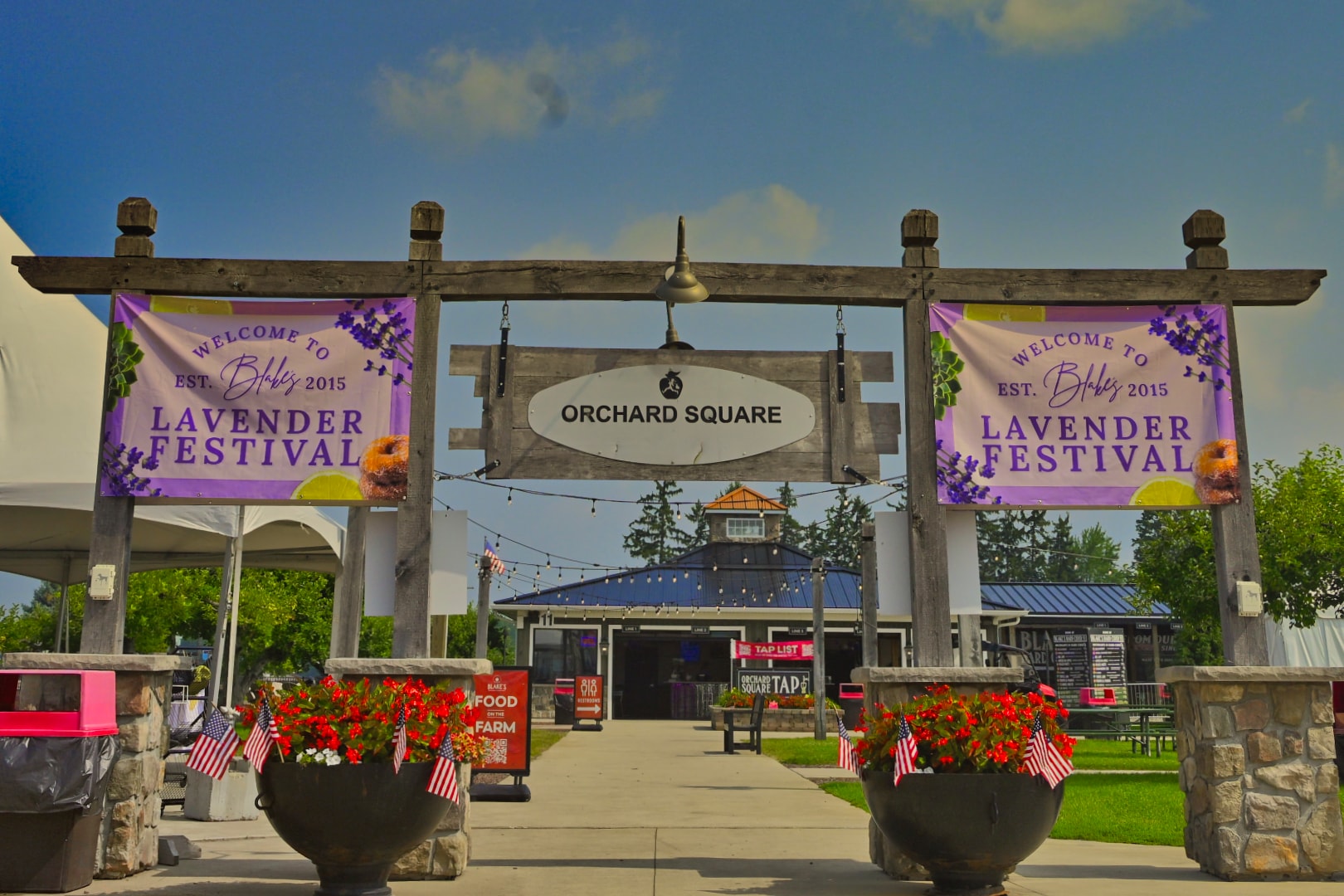 Blake's Lavender Festival welcome banners flanking the Orchard Square entrance gate at Blake's Apple Orchard in Armada, Michigan — festival signage, string lights, and flowering planters with American flags — Michigan event photography by Atelier Lens LLC