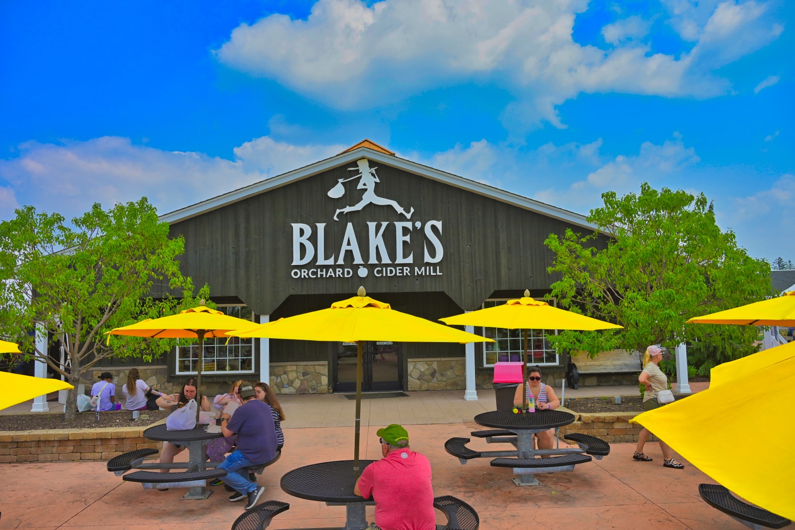 Blake's Orchard & Cider Mill main building exterior with bold white logo signage, yellow patio umbrellas, and outdoor seating area filled with guests — commercial architecture photography at Blake's Apple Orchard, Armada, Michigan by Atelier Lens LLC