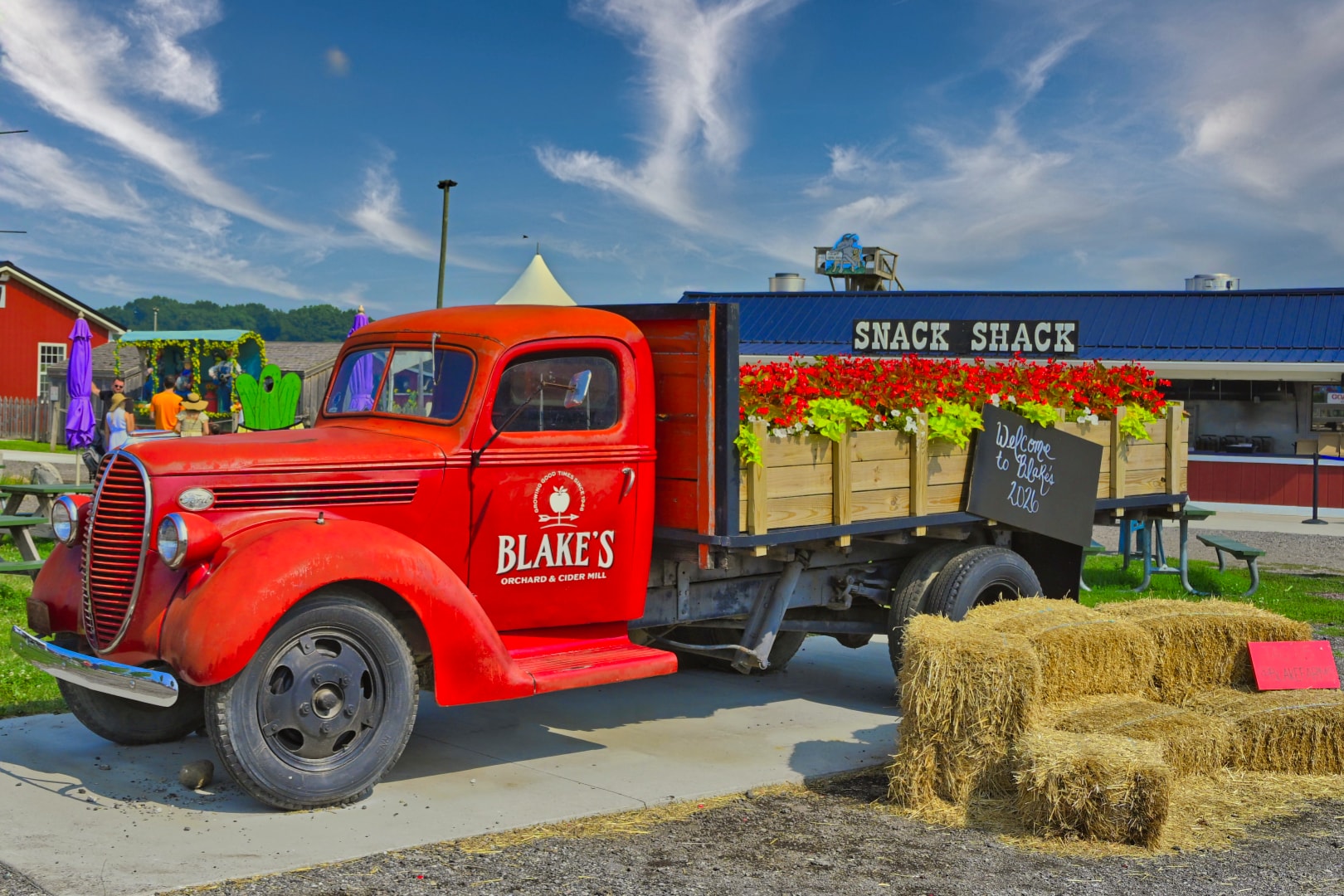 Vintage red Blake's Orchard & Cider Mill truck loaded with flower planters and a Welcome to Blake's 2020 chalkboard sign, parked in front of the Snack Shack building — commercial property photography, Armada, Michigan by Atelier Lens LLC